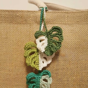 Può includere: Tre ornamenti a forma di foglia di monstera all'uncinetto in tonalità di verde e bianco sono appesi a una borsa di iuta. Le foglie sono dettagliate con un'opera traforata e la borsa ha un aspetto naturale e strutturato. Le foglie sono attaccate con un cordoncino verde e bianco.