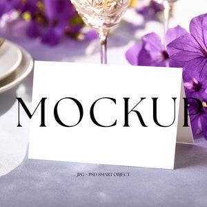 May include: A white folded card with the word "MOCKUP" in black text, set on a light purple tablecloth. Purple flowers and a crystal wine glass are in the background, creating an elegant setting. The card is a PSD smart object.
