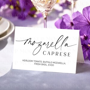 May include: A white card with the words "Mozzarella Caprese" in elegant black script. The ingredients, including heirloom tomato and fresh basil, are listed below. The card is on a table with purple flowers and a wine glass in the background.