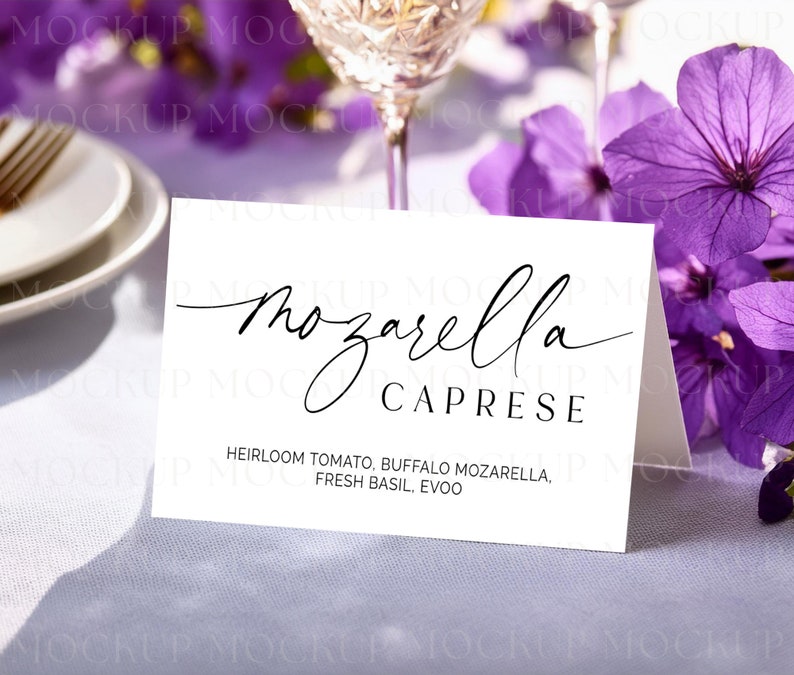 May include: A white card with the words "Mozzarella Caprese" in elegant black script. The ingredients, including heirloom tomato and fresh basil, are listed below. The card is on a table with purple flowers and a wine glass in the background.