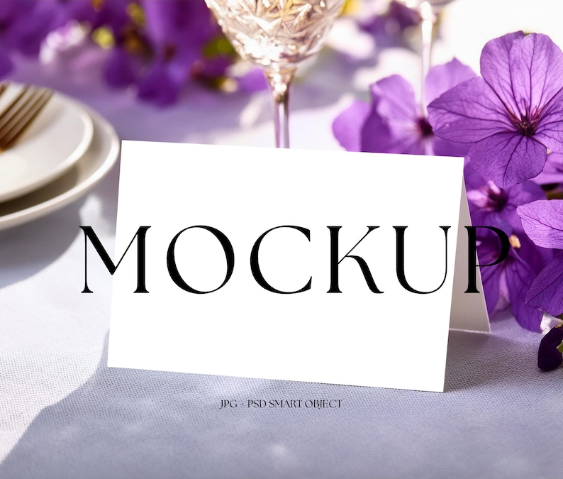 May include: A white folded card with the word "MOCKUP" in black text, set on a light purple tablecloth. Purple flowers and a crystal wine glass are in the background. The card is a mockup for a design.