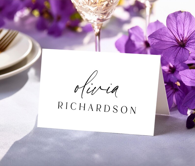 May include: A white place card with the name "olivia" in cursive and the last name "RICHARDSON" in a serif font. The card is on a white surface, with purple flowers and a wine glass in the background.