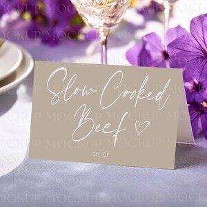 May include: A tan place card with the words "Slow Cooked Beef" in elegant white script, with a heart symbol. The card is on a white tablecloth, with purple flowers and a wine glass in the background.