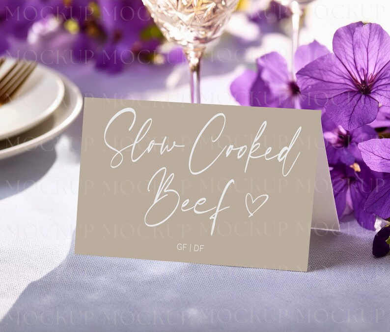 May include: A tan place card with the words "Slow Cooked Beef" in elegant white script, with a heart symbol. The card is on a white tablecloth, with purple flowers and a wine glass in the background.