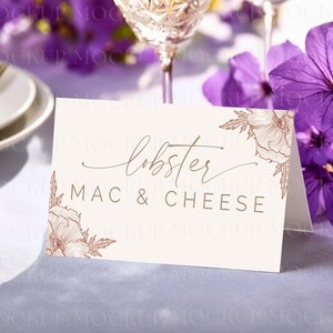May include: A cream-colored tent card with the words "lobster MAC & CHEESE" in brown script and block letters. Floral line art in brown accents the corners. The card is on a white tablecloth, with purple flowers and a wine glass in the background.