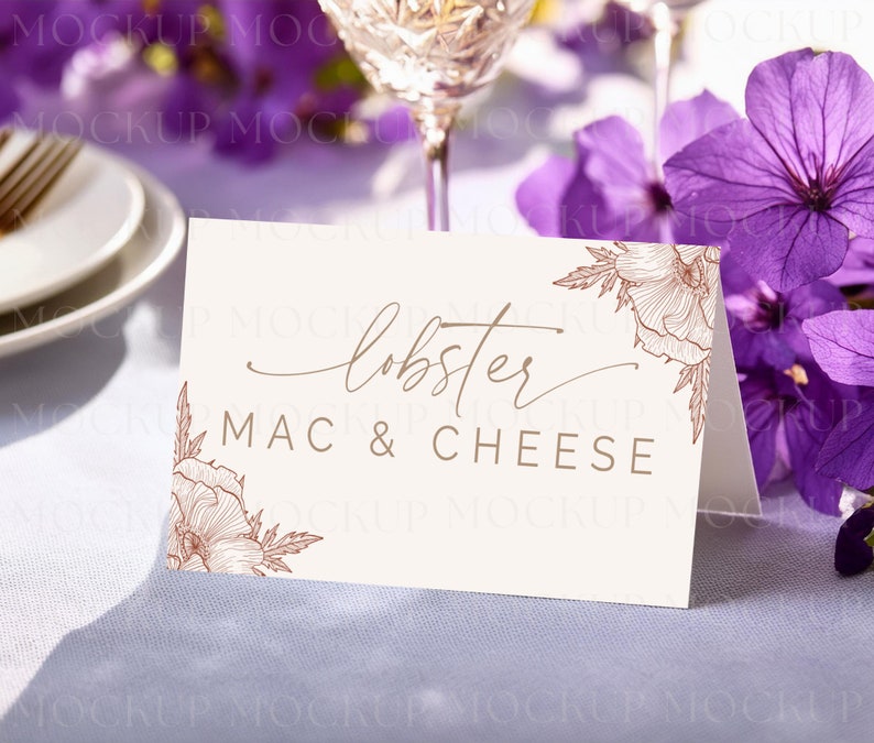 May include: A cream-colored tent card with the words "lobster MAC & CHEESE" in brown script and block letters. Floral line art in brown accents the corners. The card is on a white tablecloth, with purple flowers and a wine glass in the background.