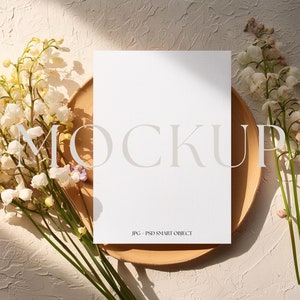 May include: A white card mockup with the word "MOCKUP" in a light gray font on a beige background. The card is on a wooden plate with white flowers on either side.