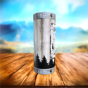 May include: A cylindrical tumbler with a rustic wood-grain design and a clear lid. The tumbler features a label with the word "CAMP" and other text, along with a silhouette of pine trees. The tumbler is designed for outdoor use.