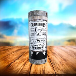 May include: A cylindrical tumbler with a rustic wood barrel design. The tumbler features the text "CABIN RULES" with outdoor-themed illustrations and phrases like "Enjoy the Outdoors". The top is clear.