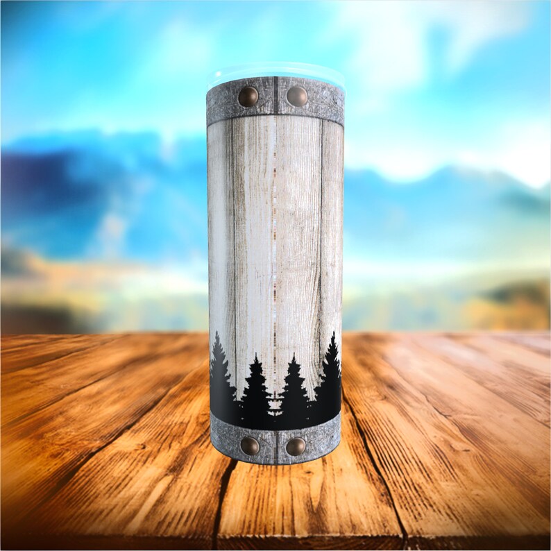 May include: A tall, cylindrical tumbler with a rustic design. The tumbler has a wood-grain pattern with a black silhouette of pine trees at the base. Metal bands with rivets encircle the top and bottom, and a clear lid is visible.