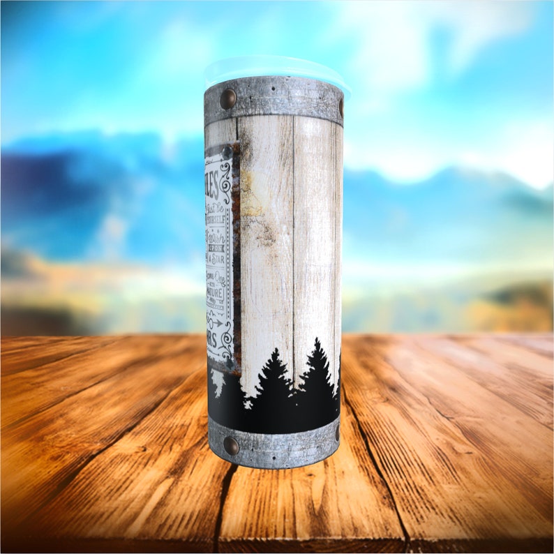 May include: A tall, cylindrical tumbler with a rustic design. The tumbler features a white wooden panel with a black silhouette of pine trees at the bottom. The design includes text and is accented with metal bands. The tumbler has a clear plastic lid.