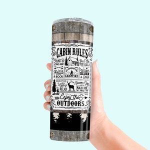 May include: A stainless steel tumbler with a wooden texture design and the words "CABIN RULES" in black and white. The tumbler features cabin-themed illustrations and phrases like "Enjoy the Outdoors."