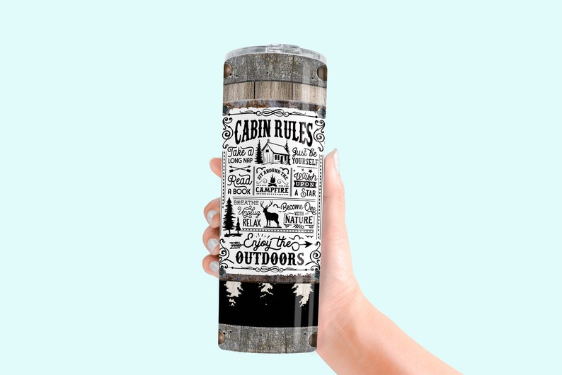 May include: A stainless steel tumbler with a wooden texture design and the words "CABIN RULES" in black and white. The tumbler features cabin-themed illustrations and phrases like "Enjoy the Outdoors."