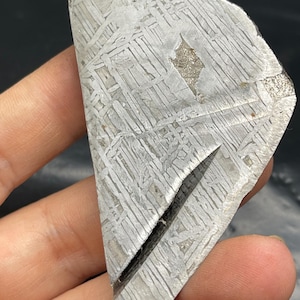 May include: A triangular-shaped, metallic meteorite fragment with a distinctive Widmanstätten pattern of intersecting lines. The fragment has a dark, irregular edge and a visible crack. The surface displays a mix of silver and gray tones.
