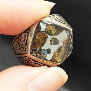 May include: A silver ring featuring a square-shaped stone set in the centre. The stone is clear and contains small pieces of brown and gold material. The ring is decorated with intricate designs around the stone.