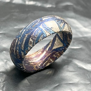 May include: A ring featuring a complex, textured surface with a metallic finish. The band displays a pattern of deep blue and purple iridescent tones contrasted with silver-coloured, etched geometric shapes that wrap around the entire circumference.