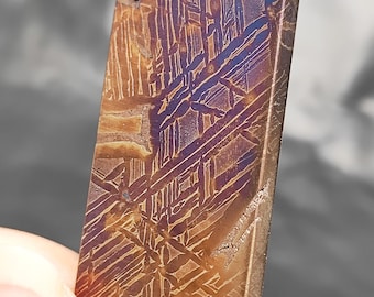 May include: A close-up of a rectangular, metallic object with a distinctive Widmanstätten pattern. The surface features a complex, cross-hatched design in shades of brown, purple, and blue. The object has a slightly rough texture and a small hole near the top.