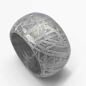 Puede incluir: Un anillo ancho y texturizado con un aspecto metálico gris. La superficie del anillo presenta un patrón único de líneas y formas que se cruzan, creando una estética industrial distintiva. Una pieza de declaración audaz.