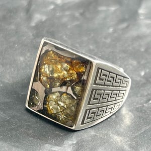 Puede incluir: Un anillo de plata con una cara cuadrada. La cara del anillo está llena de una mezcla de piedras de color oro y verde. Los lados del anillo presentan un patrón geométrico repetitivo.