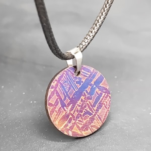 Pode incluir: Um pingente redondo com um padrão único em tons de roxo, azul e laranja. O pingente é preso a um cordão preto e a um fecho prateado. O design apresenta um padrão geométrico, criando um efeito visual marcante. Este colar é uma joia.