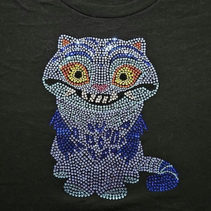 May include: Black t-shirt featuring a rhinestone-studded Cheshire Cat design. The cat has large yellow and orange eyes, a pink nose, and a wide grin. The cat's fur is primarily blue, with accents of orange and white.