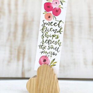 May include: A watercolor bookmark with floral accents and the text "Sweet friendships refresh the soul Proverbs 27:9." The bookmark is held by a wooden heart-shaped stand. The flowers are shades of pink and red.