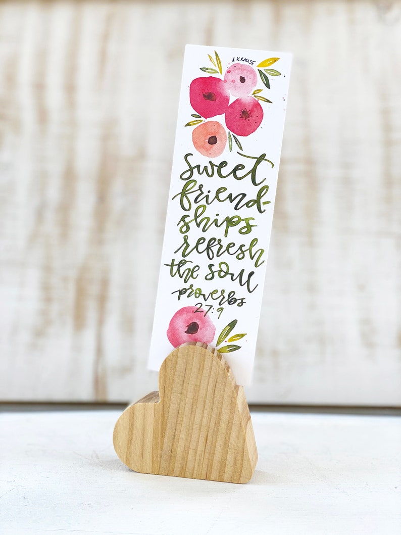 May include: A watercolor bookmark with floral accents and the text "Sweet friendships refresh the soul Proverbs 27:9." The bookmark is held by a wooden heart-shaped stand. The flowers are shades of pink and red.