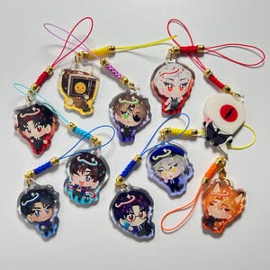 Puede incluir: Una colección de nueve dijes de acrílico con varios personajes de estilo anime, incluyendo una cabeza de televisión, un cíclope y figuras con halos. Cada dije está unido a un cordón trenzado de colores con un broche de metal dorado.
