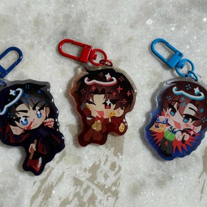 Puede incluir: Tres llaveros de acrílico con personajes de estilo anime. El llavero de la izquierda tiene un cierre azul, el central uno rojo y el de la derecha uno azul. Cada dije muestra un personaje único con colores vibrantes y rasgos faciales expresivos.