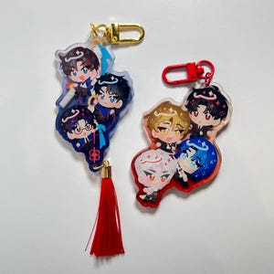 Puede incluir: Dos llaveros de acrílico con personajes de anime estilo chibi. El llavero izquierdo tiene un cierre dorado y una borla roja, mostrando tres figuras de cabello oscuro. El llavero derecho tiene un cierre rojo y tres figuras de colores variados.