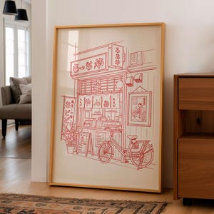 May include: A framed print featuring a red line drawing of a Japanese ramen shop with a bicycle parked in front. The shop has a sign in Japanese and a small menu board. The artwork is in a light wood frame.