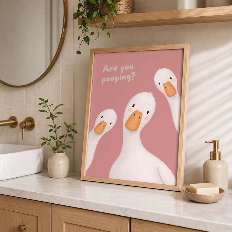 May include: A framed wall art print featuring three white geese with orange beaks on a dusty pink background. The text "Are you pooping?" is printed in white in the upper left corner. The geese have simple black eyes and pink cheek spots.