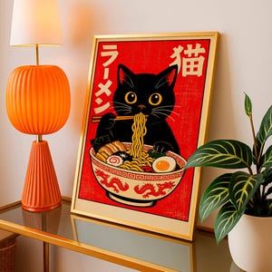 Puede incluir: Un cuadro enmarcado con un gato negro comiendo ramen. La obra, enmarcada en dorado, se encuentra sobre un fondo rojo con caracteres japoneses. Est&aacute; colocado sobre una mesa con tapa de cristal, junto a una l&aacute;mpara naranja y una planta en maceta.