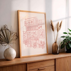 May include: Red line art print of a Japanese ramen shop with a bicycle parked out front. The shop has hanging lanterns and signage in Japanese. The artwork is framed in a light wood frame and displayed on a wooden surface.