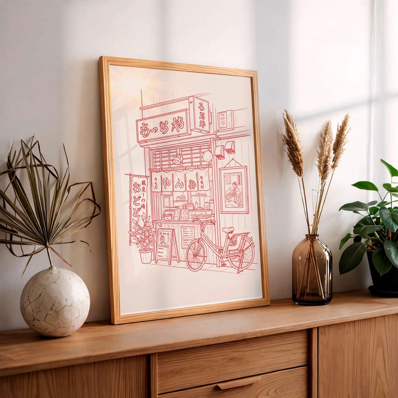 May include: Red line art print of a Japanese ramen shop with a bicycle parked out front. The shop has hanging lanterns and signage in Japanese. The artwork is framed in a light wood frame and displayed on a wooden surface.