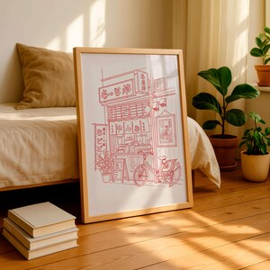 May include: A framed print featuring a red line drawing of a Japanese ramen shop with a bicycle parked in front. The print is in a light wood frame and is leaning against a bed. A stack of books sits on the floor.
