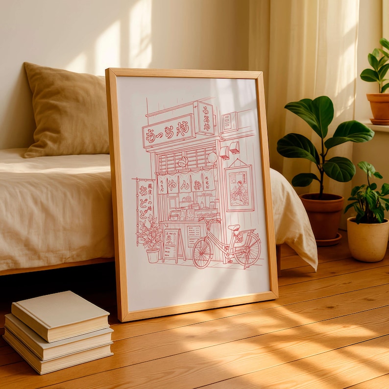 May include: A framed print featuring a red line drawing of a Japanese ramen shop with a bicycle parked in front. The print is in a light wood frame and is leaning against a bed. A stack of books sits on the floor.