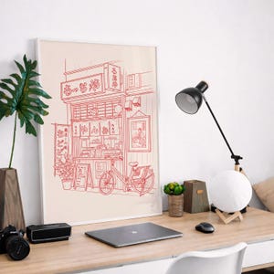 May include: A framed red and white illustration of a Japanese noodle shop with a bicycle parked in front. The shop's sign features Japanese characters. A black desk lamp, a camera, and a laptop are on the wooden desk.