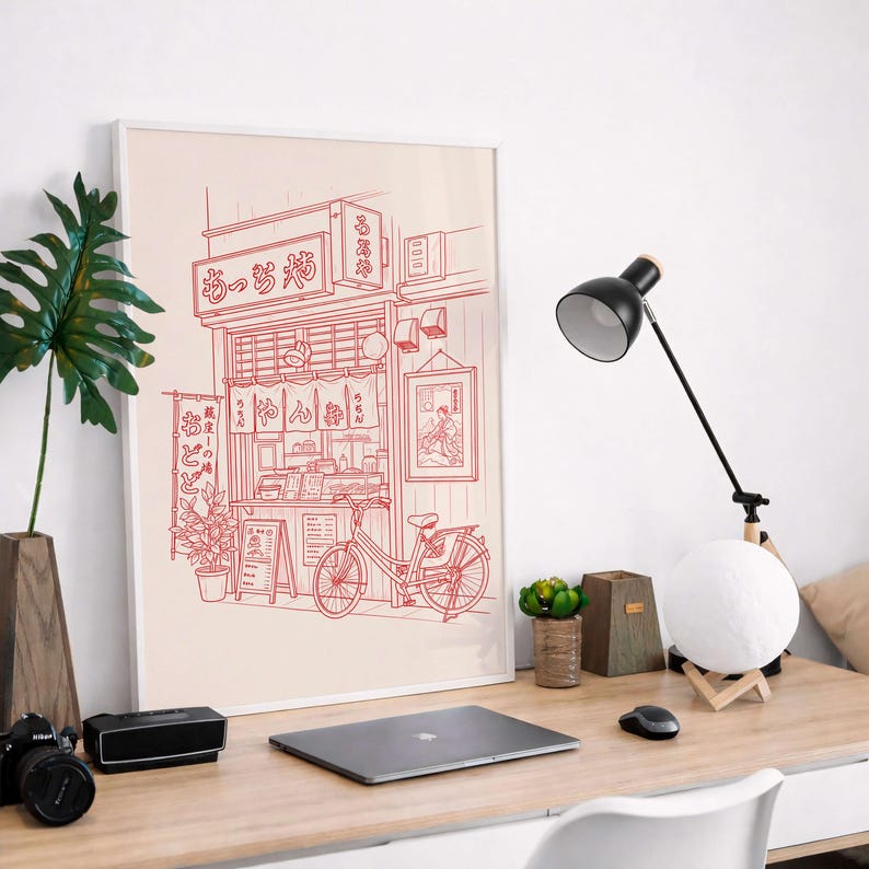 May include: A framed red and white illustration of a Japanese noodle shop with a bicycle parked in front. The shop's sign features Japanese characters. A black desk lamp, a camera, and a laptop are on the wooden desk.