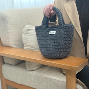 May include: A dark gray crocheted tote bag with a small rectangular label that reads "bonjour". The bag has a rounded shape with two handles. The bag is sitting on a wooden armrest.