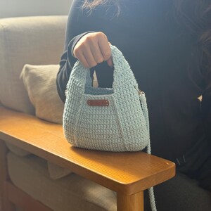 May include: A light blue crochet handbag with a short handle and a detachable shoulder strap. The bag has a rectangular shape with a small brown label. The bag is sitting on a wooden bench.