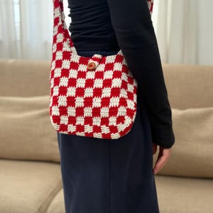Puede incluir: Un bolso de hombro de crochet a cuadros rojos y blancos con cierre de botón de madera. El bolso tiene una correa larga y está hecho de hilo suave y texturizado. Un accesorio elegante.