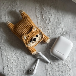 Puede incluir: Una funda de ganchillo con forma de perro Shiba Inu marrón y crema para auriculares inalámbricos, con un estuche de carga blanco y auriculares blancos. La funda está sobre una superficie blanca texturizada.