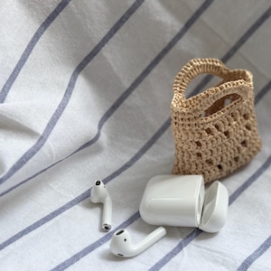 May include: White wireless earbuds and their charging case on a striped blue and white fabric with a small woven straw bag.