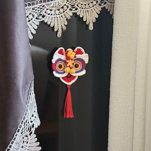 Puede incluir: Cabeza de baile del león hecha a mano en ganchillo, roja, blanca, azul y amarilla, con una borla roja. El artículo decorativo es probablemente un imán o adorno, posiblemente para una celebración cultural.
