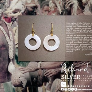 May include: A pair of white, circular earrings with gold-colored hooks. The earrings are displayed on a brown background with text that reads "Richard Anton SILVER HANDCRAFTED JEWELRY DESIGN & CREATIONS".