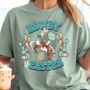May include: A light green t-shirt featuring a graphic of a bunny riding a horse with the text "Howdy Easter" in a turquoise circle. The graphic also includes cacti and Easter eggs.