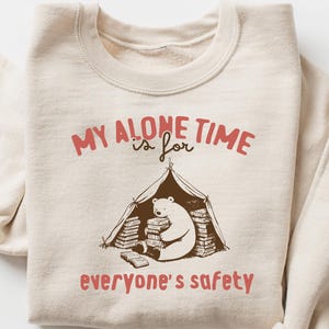 以下が含まれることがあります： ベージュのトレーナーに、茶色と赤の文字で「My alone time is for everyone's safety」と書かれています。テキストの下には、テントの中で本を読んでいるクマの漫画のイラストがあります。
