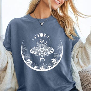 May include: A blue t-shirt featuring a white graphic of a moth, moon phases, and mushrooms. The shirt is part of a casual clothing collection. The text "Comfy Gift Shop" is visible in the bottom right corner.