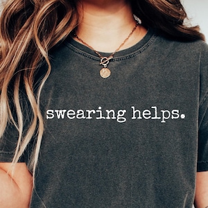May include: A dark gray t-shirt with the text "swearing helps." printed in white. The shirt is worn by a person with long brown hair and is paired with blue jeans and white sneakers.